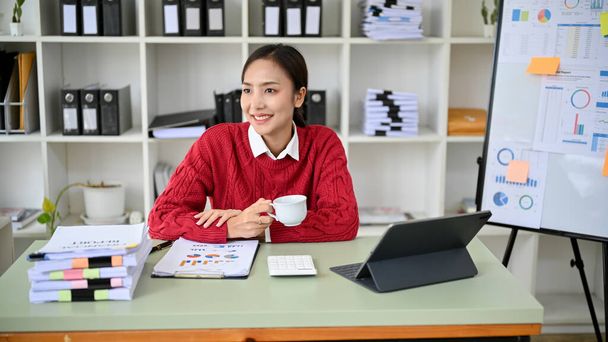 Приваблива молода азіатська бізнесменка або жінка-фінансовий аналітик, яка має ранкову каву за своїм столом, відчуває себе щасливим, посміхається і дивиться на вид з вікна.. - Фото, зображення