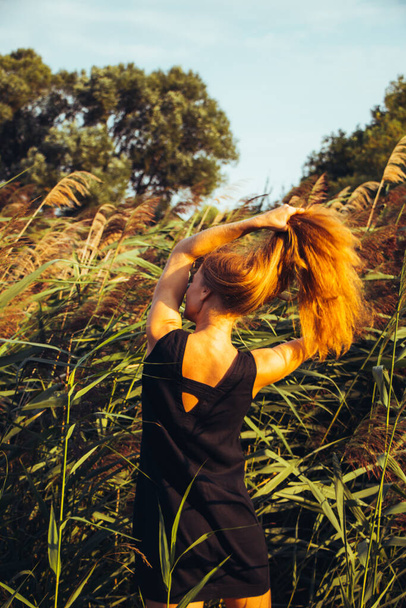 portrait of young beautiful girl in black dress with long red hair dancing smiling against green reeds. Attractive woman posing outdoors in nature, selective focus - 写真・画像
