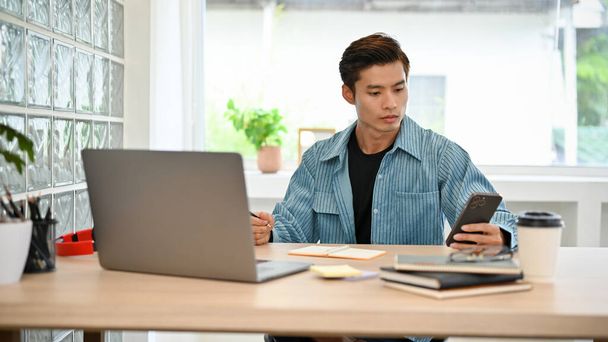 Smart young Asian businessman or male freelancer in casual outfit using his smartphone while working on his tasks at his workplace. - Photo, Image