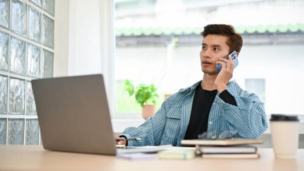 Professionnel jeune Asiatique mâle employé de bureau en vêtements décontractés parler au téléphone avec son client tout en travaillant dans le bureau. - Photo, image