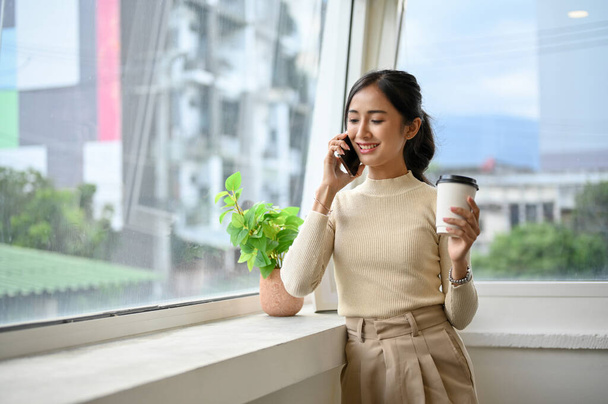 Ελκυστική νεαρή Ασιάτισσα υπάλληλος γραφείου μιλάει στο τηλέφωνο ή τηλεφωνεί στο αγόρι της κατά τη διάρκεια ενός διαλείμματος για καφέ.. - Φωτογραφία, εικόνα