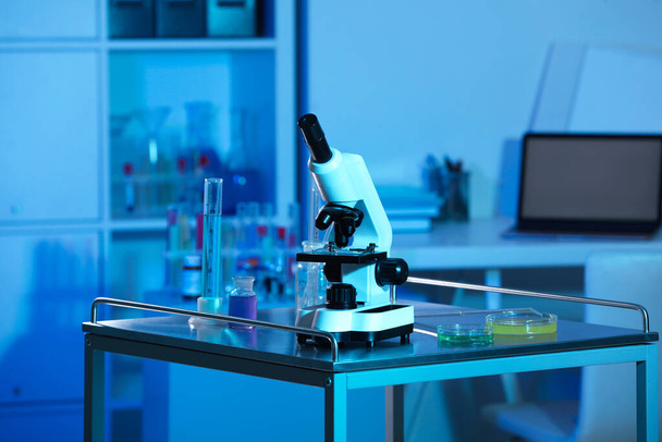 Modern microscope and glassware with colorful liquids on metal table in laboratory, toned in blue - Fotó, kép