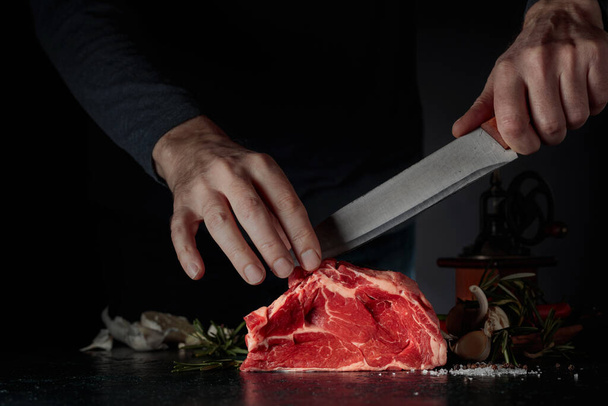 Chef cortando carne cruda de res. Sobre una mesa de carne con romero, ajo, sal y pimienta. - Foto, Imagen