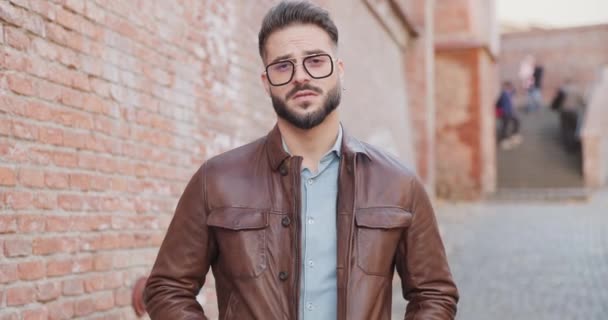 handsome cool man fixing eyeglasses, crossing arms and smiling in front of bricks wall outdoor in a medieval town from Romania - Footage, Video