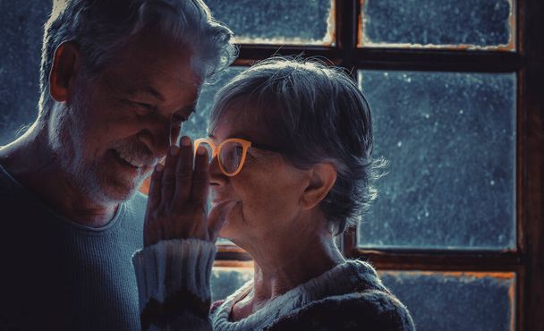Старшая женщина шепчет своему зрелому мужу дома. Пожилая пара влюблённая и влюблённая. Раскрывать секреты. Портрет пожилого человека на пенсии дома. Зимний снег за окнами сзади - Фото, изображение