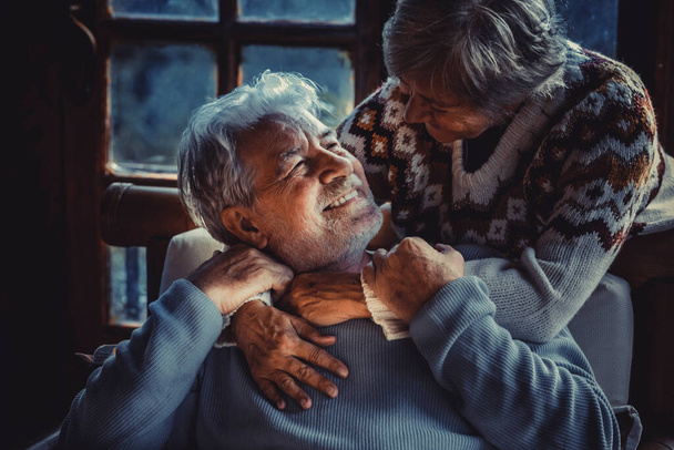 Πορτραίτο ενός ζευγαριού ηλικιωμένων ερωτευμένων στο σπίτι τους το βράδυ σκοτεινό φως. Ο γέρος χαμογελάει στην ώριμη γυναίκα του αγκαλιάζοντας και απολαμβάνοντας τη σχέση. Χειμερινό σπίτι. Η ζωή των ηλικιωμένων - Φωτογραφία, εικόνα