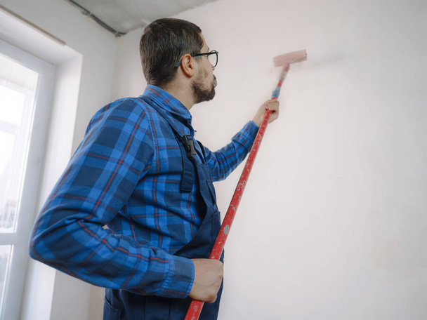 Молодой человек в синем рабочем костюме держит ролик с краской на фоне белой стены. Аксессуары для ремонта жилых помещений. Ремонт дома концепции - Фото, изображение