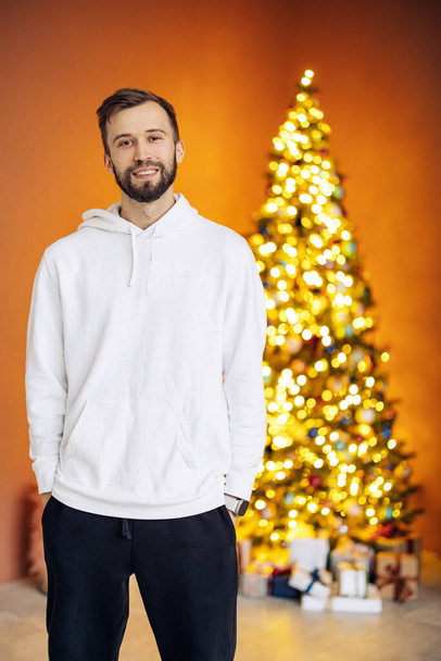 Young smiling man in hoody stands against background of decorated glowing Christmas tree and gifts. Christmas concept. Orange background. - Photo, Image