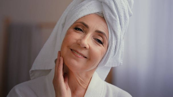 Close up female portrait happy 50s middle-aged lady 60s mature woman touching facial skin looking at camera with smile touch face pampering wears towel on head enjoy cosmetics and plastic surgery - Photo, Image
