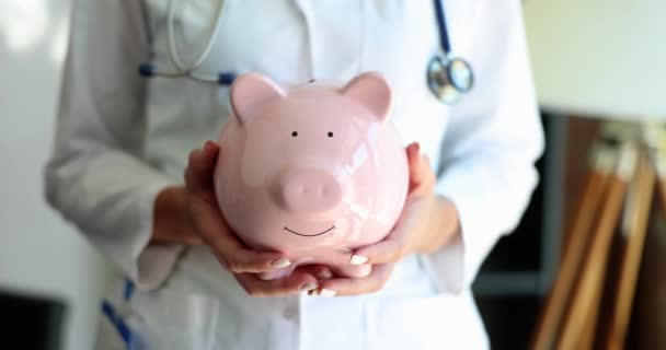 Γυναίκα γιατρός κρατάει κουμπαρά στην κλινική. Εξοικονόμηση χρημάτων από τα έξοδα λογιστικής κλινικής ιατρικής περίθαλψης - Πλάνα, βίντεο