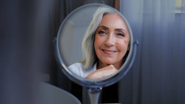 Χαμογελώντας όμορφο θηλυκό πρόσωπο 50s μεσήλικες ώριμη γυναίκα απολαμβάνουν το δέρμα του προσώπου κοιτάζοντας καθρέφτη περιποίησης αντανάκλαση. Παλιά γήρανση ξηρή περιποίηση της επιδερμίδας διαδικασίες ομορφιάς καλλυντικά αντι ηλικία πλαστική χειρουργική επέμβαση - Φωτογραφία, εικόνα