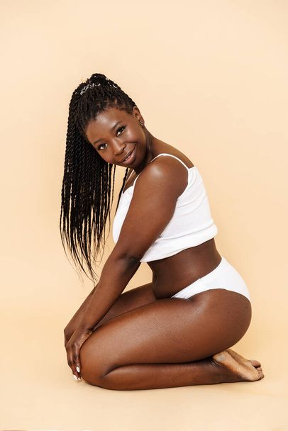 Black young woman wearing white underwear sitting on floor isolated over beige background - Photo, image