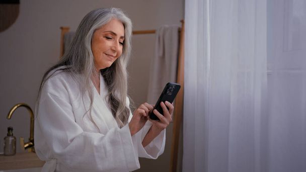 60 'ların Kafkasyalı yaşlı olgun kadın bornozla banyoda durmuş cep telefonundaki nemlendirici cilt bakımını yenilemek için makyaj kremi sipariş eden telefon rehberine bakıyor. - Fotoğraf, Görsel