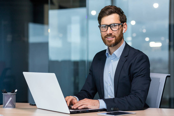 Portrait of mature successful and happy businessman, senior man with beard smiling and looking at camera, investor working with laptop inside office in business suit. - Foto, afbeelding
