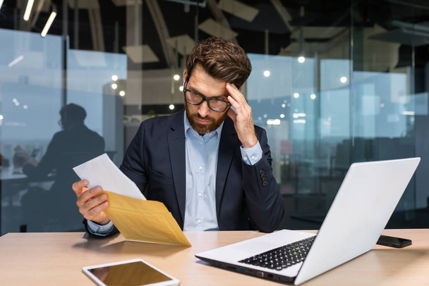 Sad and upset mature businessman received an envelope by mail with a bad news notification letter, boss in business suit working inside office using laptop wearing glasses and beard. - Foto, Imagem