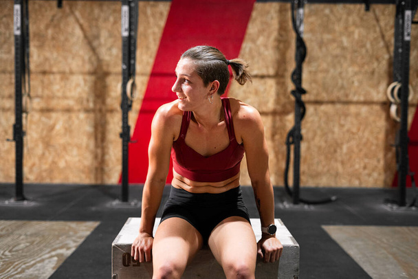 tired caucasian girl smiling after doing a lot of effort on a wooden box in a gym - Crossfit concept - Photo, Image