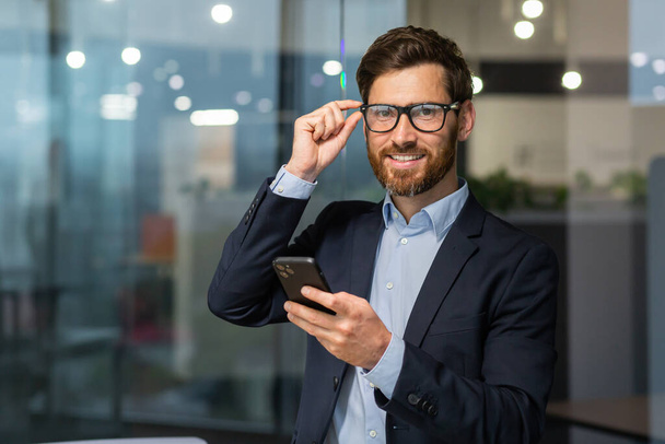 Portrait successful boss with a phone, a man in glasses and a business suit is looking at the camera, an investor is holding a smartphone in hands, reading a message and using an online application. - Fotó, kép