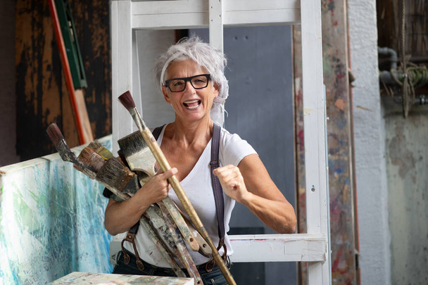 ouder grijs haar volwassen gelukkig kunstenaar vrouw met bril en grote penselen maken vuist power gebaar, kopieer ruimte - Foto, afbeelding