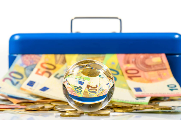 billetes y monedas de euros que yacen sobre la mesa y que sobresalen de la caja azul con reflejo en la bola de vidrio - Foto, Imagen
