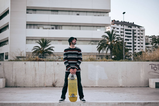 kavkazský mladý muž v pruhovaném svetru s otevřenou nohou držící skateboard s rukama na ulici - Lfestyle koncept - Fotografie, Obrázek