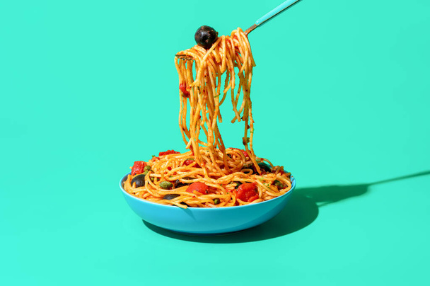 Tabaktan çatalla makarna almak, yeşil masada minimalist olmak. Vejetaryen İtalyan yemeği, spagetti puttanesca, domates sosu, siyah zeytin ve kapari.. - Fotoğraf, Görsel