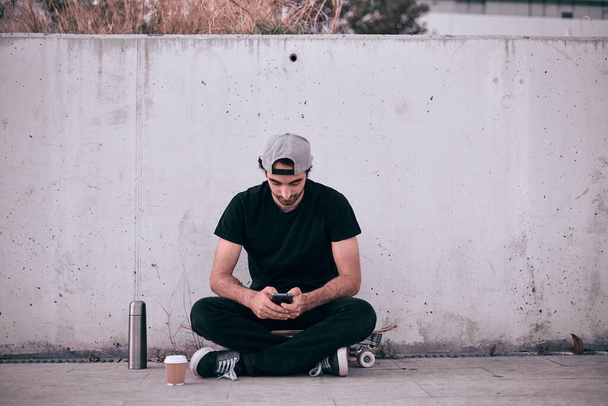 caucasian guy in gray cap and black t-shirt sitting on his skateboard calm and relaxed manipulating his smarphone while having hot coffee by the wall - Lfestyle concept - Photo, Image