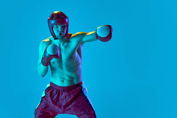 Energetic workout of professional athlete. Kickboxer in helmet and gloves training isolated over blue background in neon light. Muay thai. Sport, competition, energy, combat sports - Photo, image