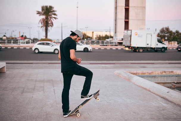 caucasian young man in black t-shirt and cap on his head standing on his skateboard in the city - Lfestyle concept - Photo, Image