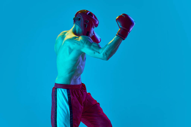 Energetic workout of professional athlete. Kickboxer in helmet and gloves training isolated over blue background in neon light. Muay thai. Sport, competition, energy, combat sports - Photo, Image