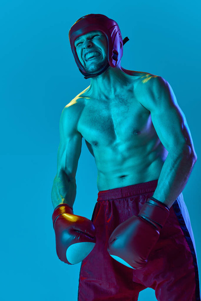 Energetic workout of professional athlete. Kickboxer in helmet and gloves training isolated over blue background in neon light. Muay thai. Sport, competition, energy, combat sports - Photo, Image