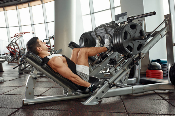 Ο άνθρωπος χρησιμοποιώντας μια μηχανή τύπου στο γυμναστήριο. Ισχυρός άνδρας κάνει μια άσκηση στα πόδια της στον προσομοιωτή. Πλάγια όψη - Φωτογραφία, εικόνα