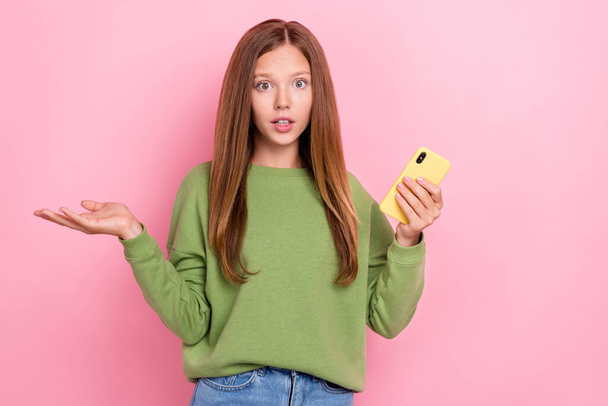 Portrait of adorable confused cute girl with long hairstyle dressed green sweatshirt hold smartphone isolated on pink color background. - 写真・画像