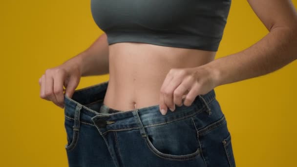 Onherkenbaar meisje vrouw toont gewichtsverlies dragen van oude jeans grote maat slanke dame check resultaat van het eten van gezonde voeding dieet fitness sport sportschool lichaam afslanken taille in studio gele achtergrond - Video