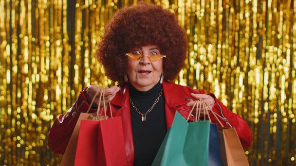 Щаслива туристична жінка з пишним волоссям, що показує торгові сумки, на свята Чорної п "ятниці усміхається знижка з рекламою, яка виглядає вражена низькими цінами. Старша стильна бабуся на блискучому фоні - Фото, зображення
