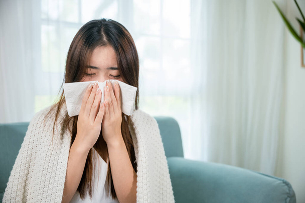 Ill Asian young woman cold covered with blanket sitting on sofa and sneeze with tissue paper at home, Female sick allergic blowing nose sneezing in tissue at home, Flu health care - Fotografie, Obrázek