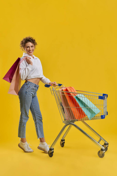 Side view of slim girl with blonde curly hair doing shopping. Pretty young woman standing on toes, driving cart, holding packages, looking at camera. Concept of urban life. - Foto, afbeelding