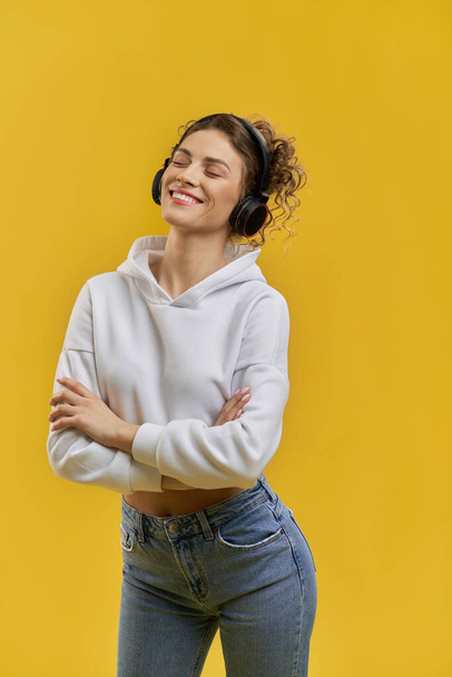 Side view of pretty slim girl standing, listening to music in earphones. Attractive young female smiling with closed eyes, crossing arms, enjoying. Concept of youth culture. - Foto, Bild
