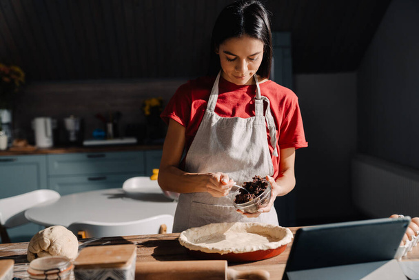 Young hispanic woman wearing apron making pie in kitchen at home - Foto, imagen