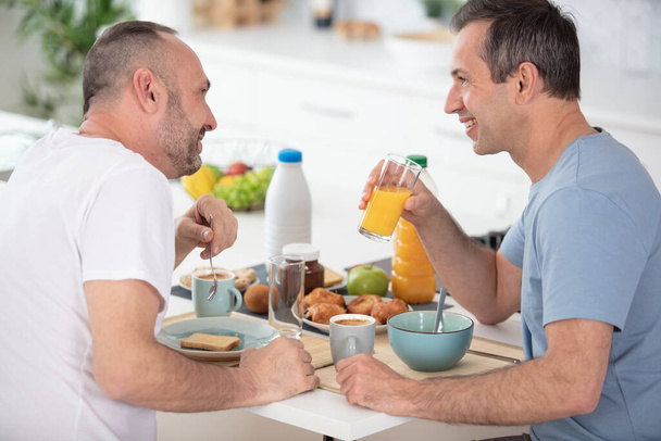 happy male partners eating breakfast and cooking in kitchen - Фото, изображение