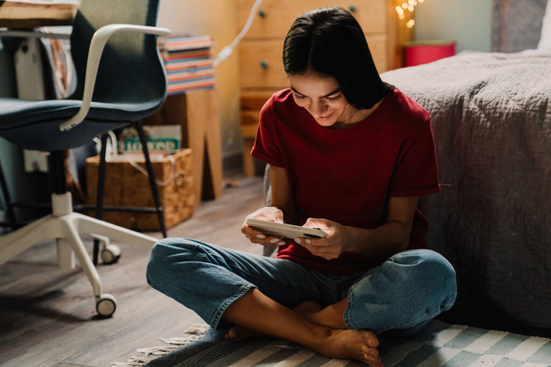 Young hispanic woman smiling and using cellphone while sitting on floor at home - Photo, Image