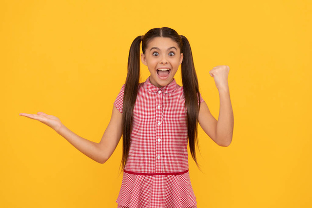 surprised teen girl presenting product on yellow background copy space celebrate success, promotion. - Foto, imagen