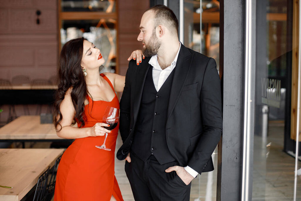 Loving couple is spending time together in modern restaurant and drinking wine. Attractive young woman in red dress and handsome man in black suit are hugging. Celebrating saint Valentines Day. - Foto, Imagen