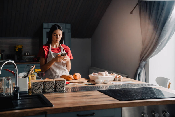 Young hispanic woman wearing apron using cellphone while cooking in kitchen at home - Фото, зображення