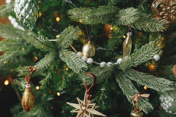 Χριστουγεννιάτικο δέντρο με vintage μπιχλιμπίδια και χρυσά φώτα κοντά. Μοντέρνα διακοσμημένα κλαδιά χριστουγεννιάτικου δέντρου με κομψά στολίδια στο εορταστικό δωμάτιο. Προετοιμασία χειμερινών διακοπών, ατμοσφαιρικός χρόνος - Φωτογραφία, εικόνα