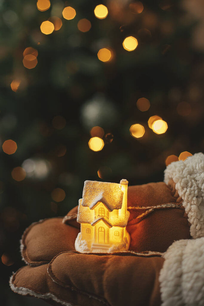 Ręce w rękawiczki trzymając mały świecący dom na tle oświetlonych świateł choinkowych. Magiczny czas zimowy, obraz atmosferyczny. Przytulny dom. Wesołych Świąt i Wesołych Świąt! - Zdjęcie, obraz