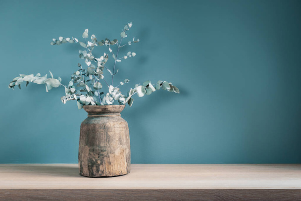 Rustic vase with dry eucalyptus leaves on wooden table against blue green wall. Home decor. - Φωτογραφία, εικόνα