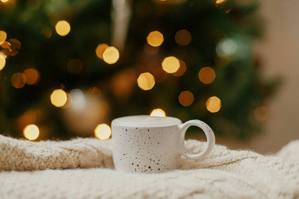 Hygge invernal atmosférico. La taza caliente del café sobre el fondo del árbol de Navidad con las luces. Acogedora casa. Taza elegante en suéter de punto contra bokeh iluminación dorada. Banner de Navidad - Foto, imagen