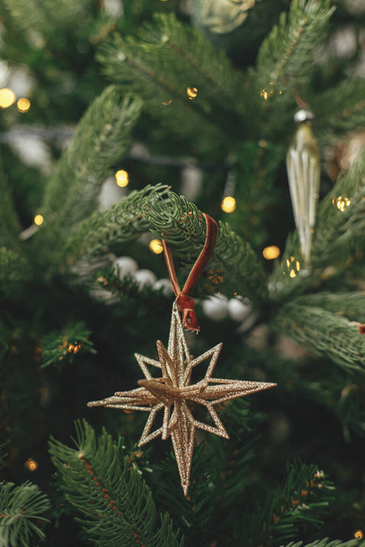 Christmas tree with vintage star and baubles with golden lights close up. Modern decorated christmas tree branches with stylish ornaments in festive room. Winter holidays, atmospheric time - Photo, Image