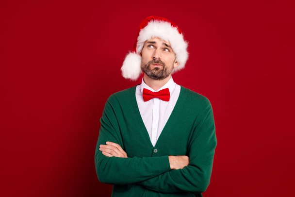 Photo of young minded grimace man elegant garment wear santa claus hat look curious up unsure folded arms idea isolated on red color background. - Φωτογραφία, εικόνα