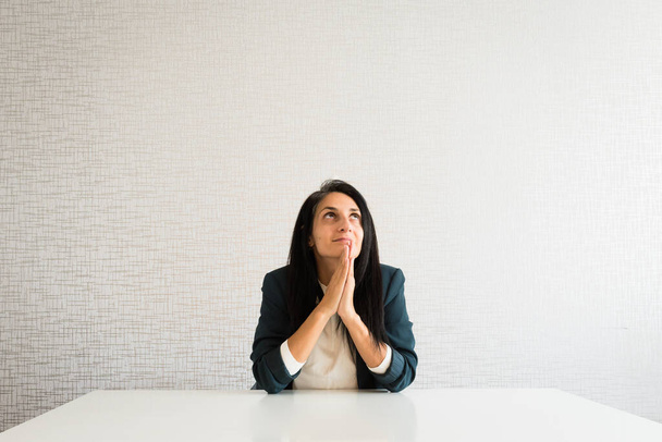 Young caucasian brunette business woman director in office pray for help hopeful in office behind desk for success and praise at work - Fotografie, Obrázek